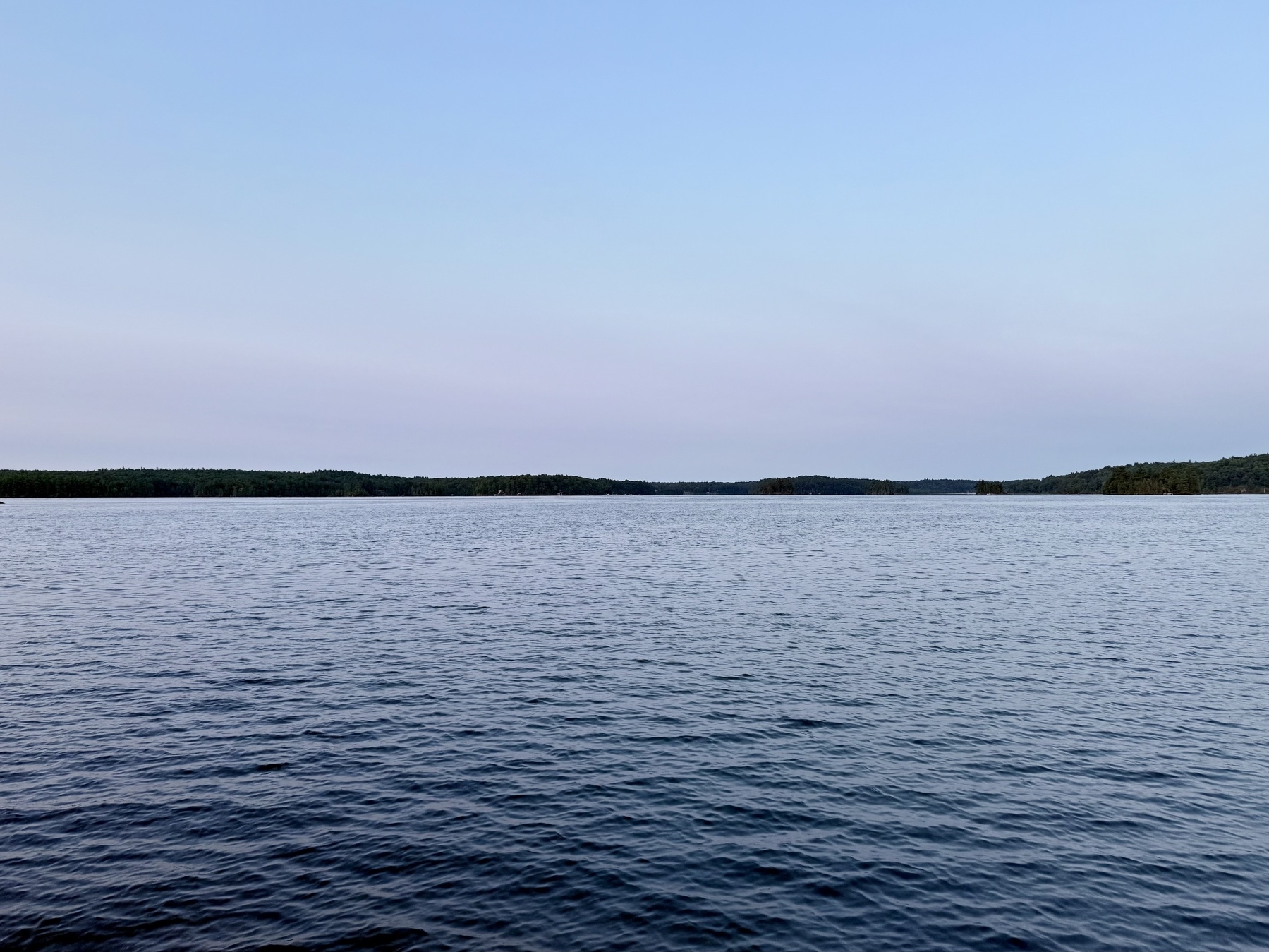 A serene body of water stretches toward a distant forested shoreline under a clear sky.