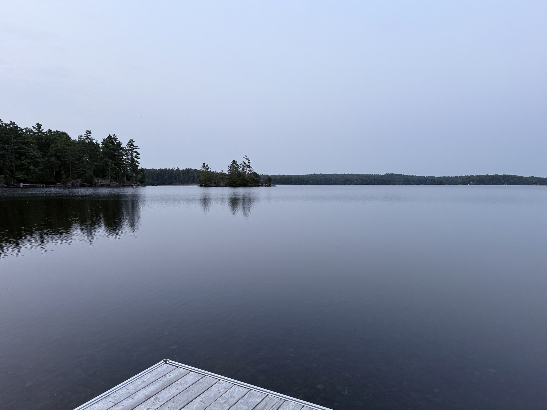 A calm lake reflects a cloudy sky, surrounded by a forested shoreline with a wooden dock in the foreground.